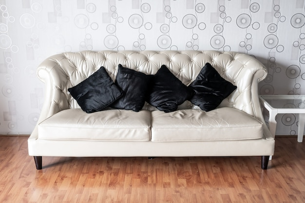 Диван кремового цвета с черными подушками на фоне узорчатых обоев.